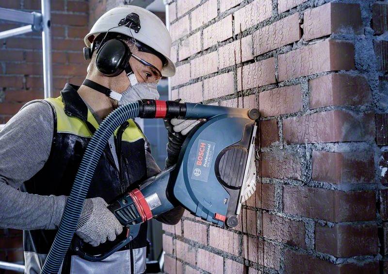 Ein Bauarbeiter mit Schutzhelm, Gehörschutz und Maske verwendet eine elektrische Säge, um Mörtelfugen in einer Ziegelwand zu schneiden.