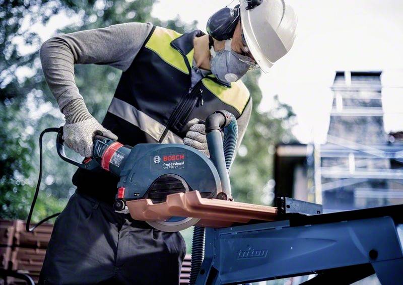Eine Person schneidet mit einer elektrischen Säge Holz, trägt Schutzkleidung und Maske. Im Hintergrund Baum und unscharfes Baugerüst.