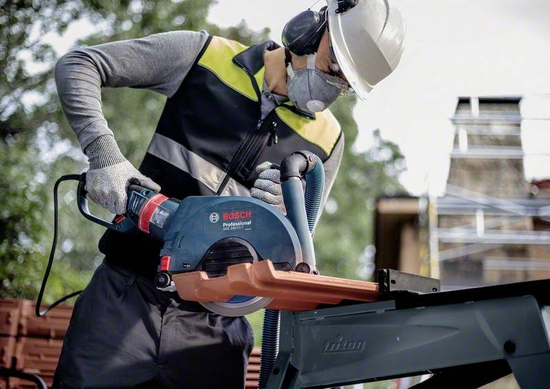 Person mit Schutzkleidung arbeitet mit einer Bosch-Handkreissäge an einem Metallstück im Freien. Hintergrund zeigt Bäume und Baugerüst.