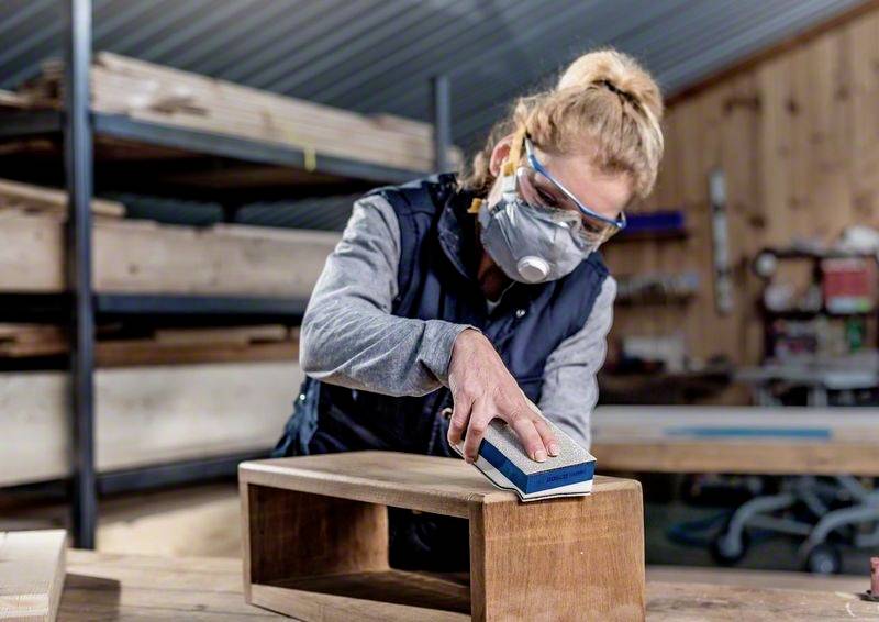 Eine Person schleift mit Schutzbrille und Atemmaske ein Holzstück in einer Werkstatt. Im Hintergrund sind Regale mit Holzmaterialien.