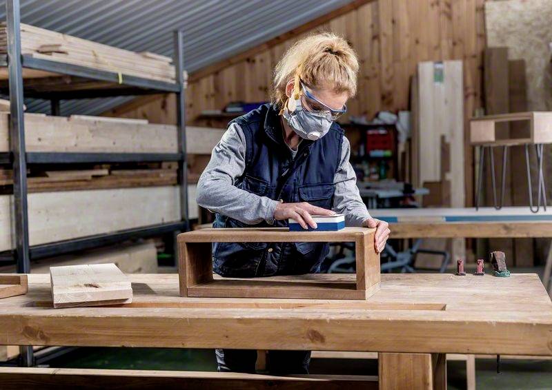 Eine Person arbeitet in einer Holzwerkstatt, schleift ein Holzstück auf einem großen Tisch. Im Hintergrund Regale mit Holzmaterialien.