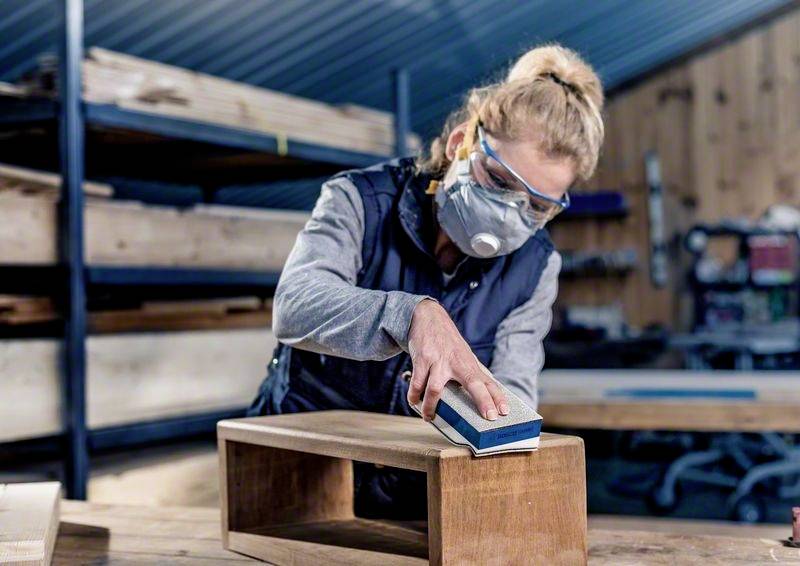 Person schleift ein Holzstück in einer Werkstatt. Trägt Schutzbrille und Atemschutzmaske. Hintergrund zeigt Holzregale und Werkzeuge.