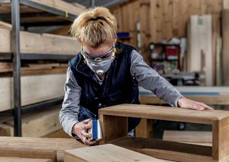 Eine Person schleift Holz in einer Werkstatt. Sie trägt Schutzbrille und Atemschutzmaske. Im Hintergrund sind Holzregale sichtbar.
