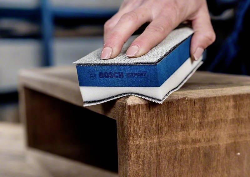 Eine Hand schleift mit einem Bosch-Schleifblock eine Holzkante. Die Oberfläche des Holzbretts wird geglättet.