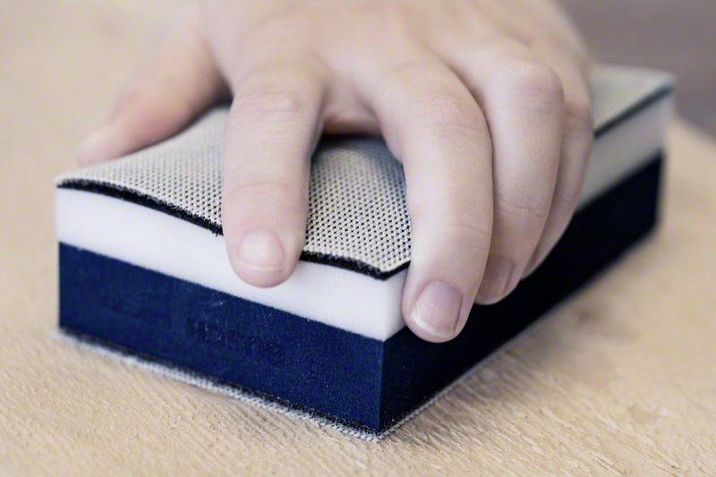 Eine Hand hält einen rechteckigen Schwamm mit einer weißen und schwarzen Schicht, auf einer Holzoberfläche.