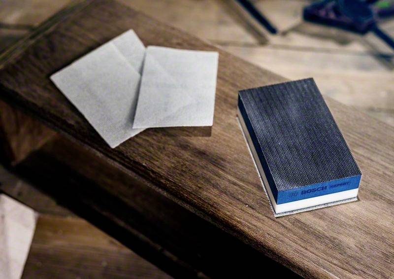 Ein Schleifblock und zwei Schleifpapiere liegen auf einem Holztisch. Im Hintergrund sind Werkzeuge zu sehen.