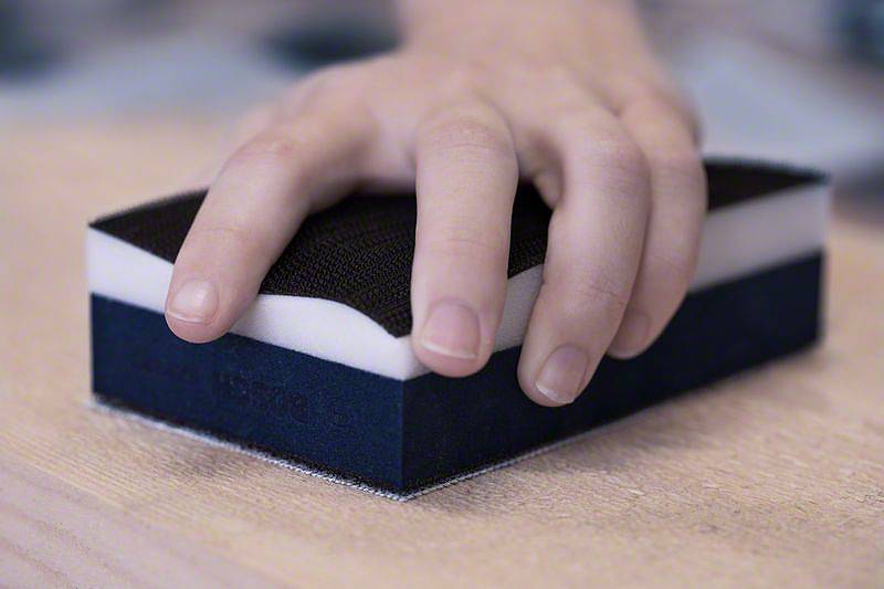 Eine Hand hält einen Schleifschwamm, der auf einer Holzoberfläche verwendet wird. Der Schwamm hat eine schwarze und weiße Schicht.