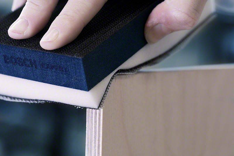 Eine Hand verwendet einen Schleifschwamm, um die Kante eines Holzbretts zu glätten, was auf eine handwerkliche Tätigkeit hindeutet.