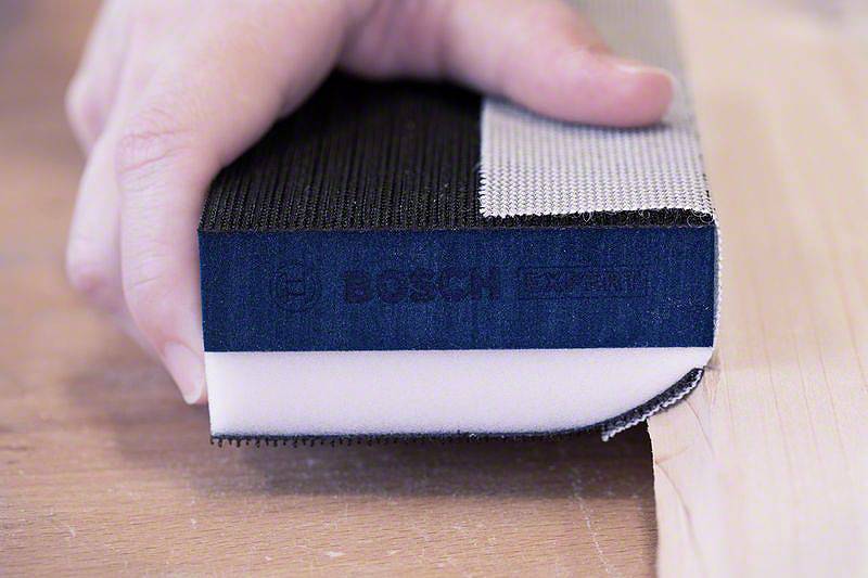 Eine Hand hält einen Schleifschwamm der Marke Bosch auf einem Holzbrett, bereit zum Schleifen.