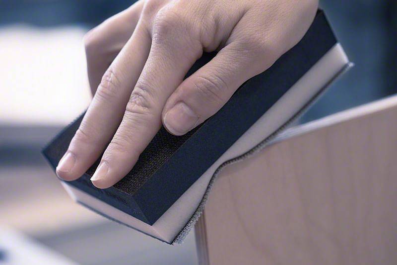 Eine Hand schleift die Kante eines Holzteils mit einem Schleifblock.