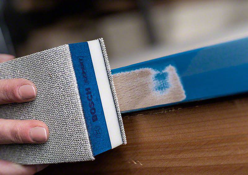 Hand schleift blaue Kante eines Holzteils mit einem Schleifblock. Holzfarbe wird sichtbar. Fokus auf Präzision und Handwerk.