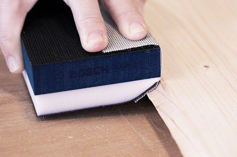 Eine Hand hält einen Schleifschwamm auf Holz, um die Oberfläche zu glätten. Der Schwamm zeigt das Logo eines bekannten Werkzeugherstellers.