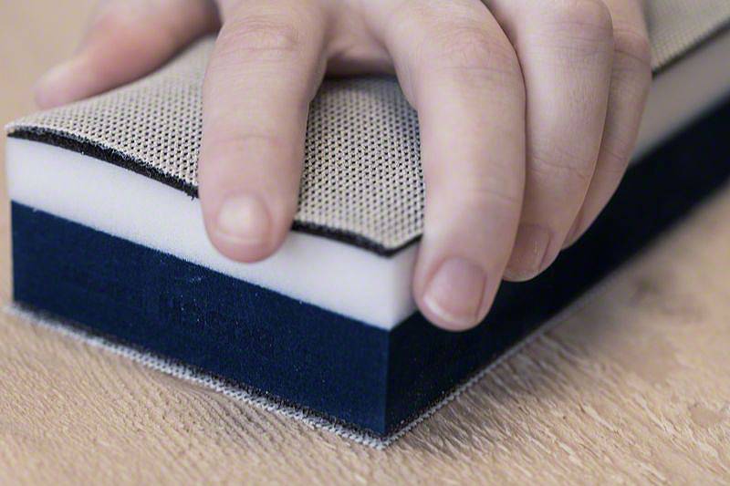 Eine Hand hält einen rechteckigen Schwamm mit weißen, grauen und blauen Schichten auf einer Holzoberfläche.