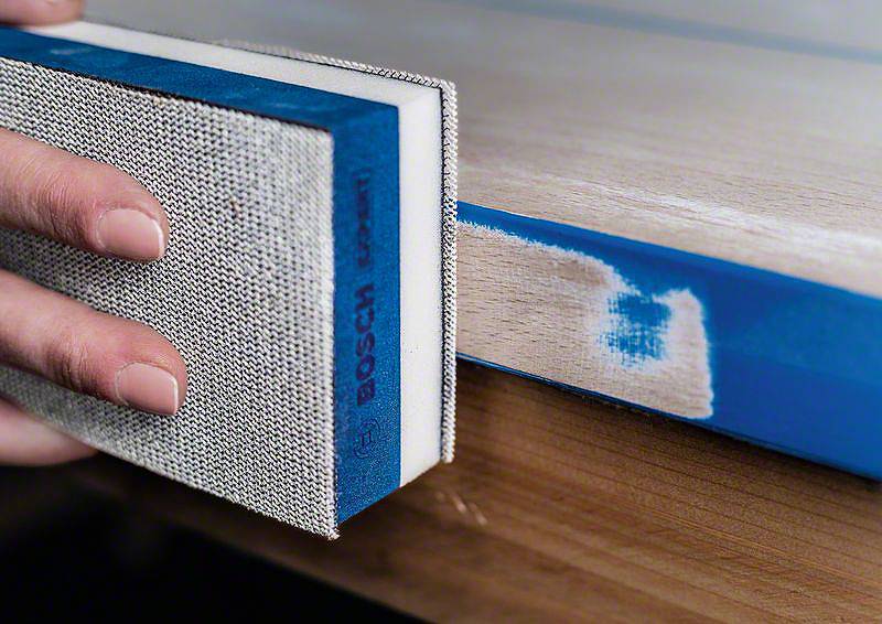 Eine Hand schleift die Kante eines Tisches mit einem blauen Schleifblock, um die Oberfläche zu glätten.