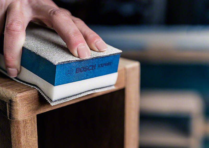 Eine Hand hält einen Schleifschwamm und schleift die Kante eines Holzmöbelstücks. Der Schwamm trägt das Logo einer bekannten Marke.