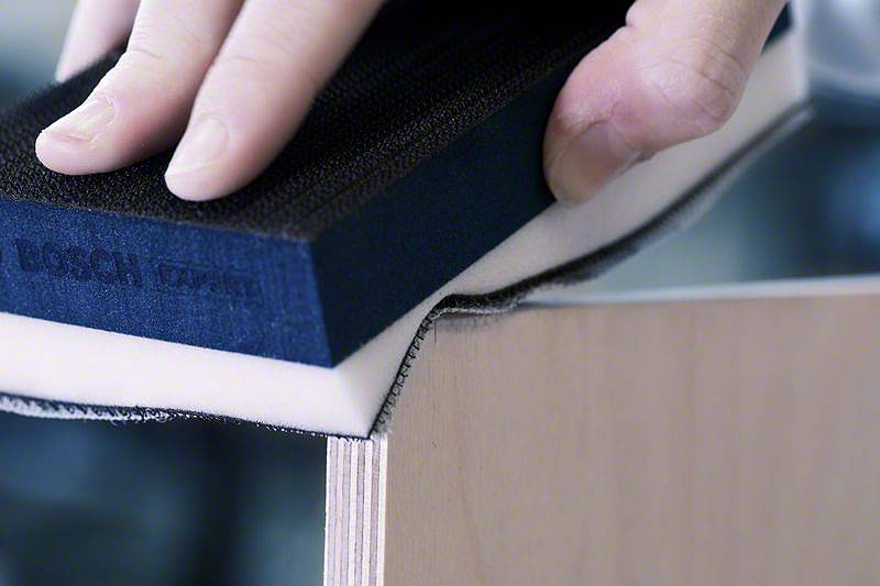 Jemand schleift die Kante eines Holzbretts mit einem Schleifblock. Fokus liegt auf der Hand und dem Schleifblock.