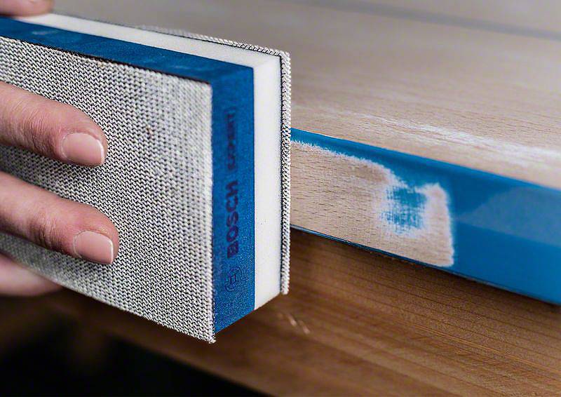 Eine Person schleift die Kante eines Holzbretts mit einem Schleifpad, um die Oberfläche zu glätten. Der Rand ist blau markiert.