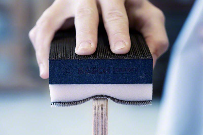 Eine Hand hält einen Bosch-Schleifschwamm, der auf einem Holzbalken eingesetzt wird. Der Schwamm ist zweifarbig: schwarz und weiß.