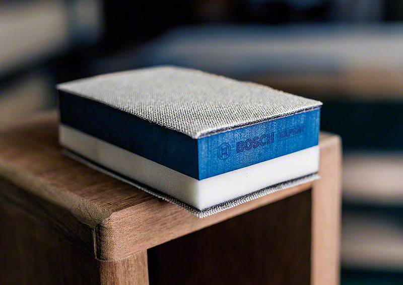 Ein blauer Bosch-Handschleifblock liegt auf einem Holztisch. Der Schleifblock hat eine grobe, graue Schleiffläche und eignet sich zum Glätten von Holzoberflächen.