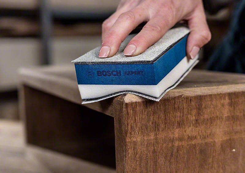 Hand schleift Holz mit einem Bosch Schleifschwamm. Der Fokus liegt auf der Präzision des Schleifens und der markanten blauen Farbe des Schwamms.