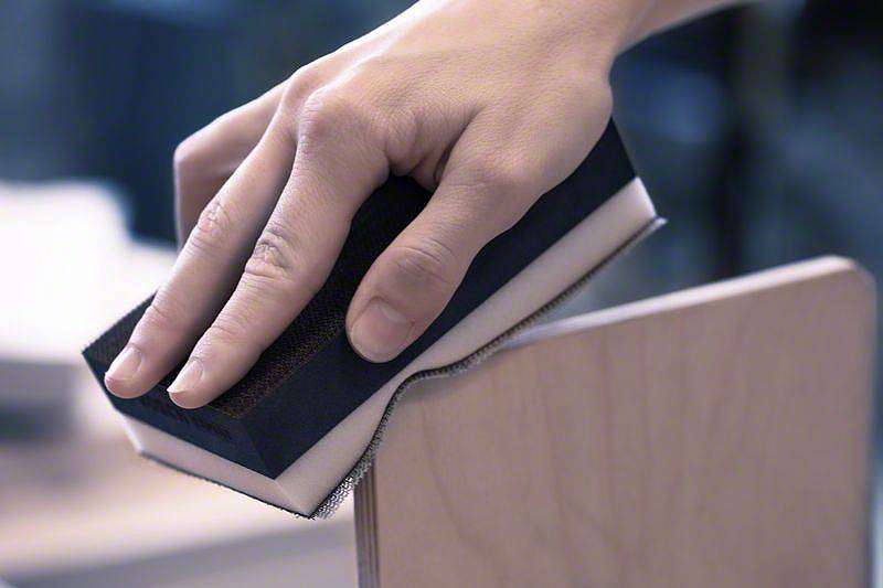 Eine Hand schleift mit einem Schleifblock die Kante eines Holzbretts.