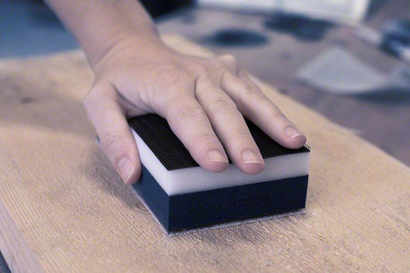 Eine Hand hält einen Schleifschwamm und bearbeitet eine Holzoberfläche. Der Schwamm hat eine schwarze Schleifseite.