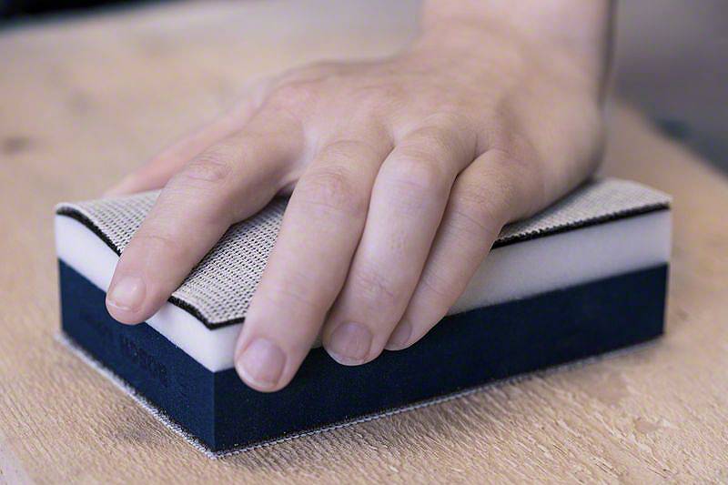 Eine Hand hält einen Schleifblock, der über eine Holzoberfläche gleitet, um diese zu bearbeiten.