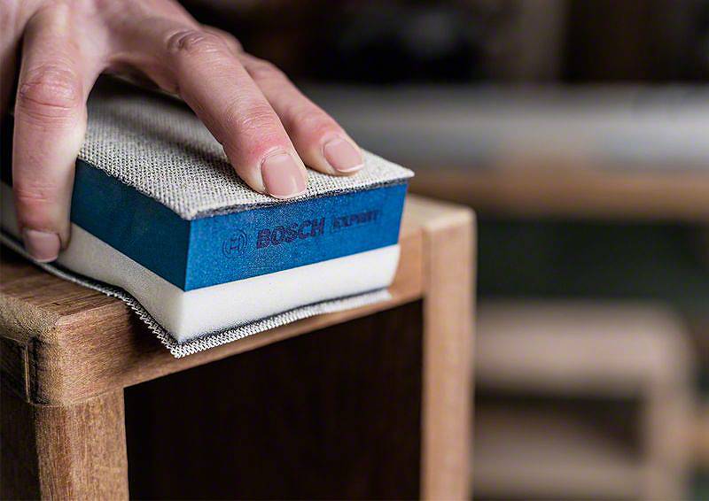 Eine Hand schleift mit einem Schleifblock eine Holzoberfläche. Der Schleifblock ist blau mit dem Aufdruck 'BOSCH Expert'.
