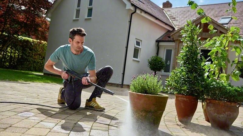 Ein Mann reinigt mit einem Hochdruckreiniger Pflastersteine vor einem Haus. Es ist sonnig, und Pflanzen stehen in Töpfen neben ihm.