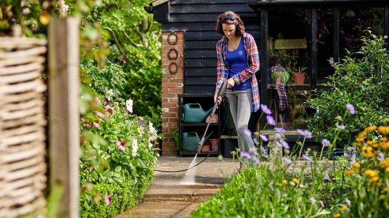 Eine Person reinigt mit einem Hochdruckreiniger einen Weg im Garten. Links und rechts blühen bunte Blumen.