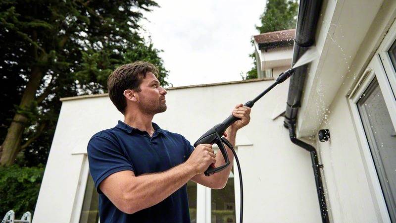 Ein Mann spritzt Wasser mit einem Hochdruckreiniger auf die Dachrinne eines Hauses, um sie zu reinigen. Im Hintergrund sind Bäume zu sehen.