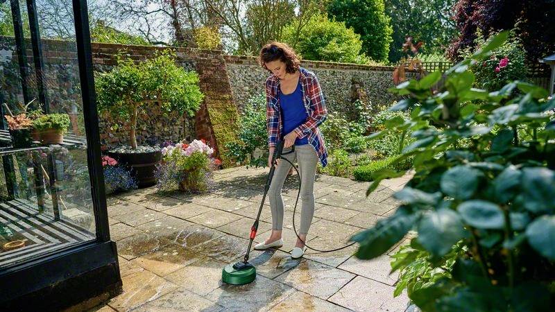 Eine Frau reinigt eine Terrasse im Garten mit einem Hochdruckreiniger. Umgeben von Pflanzen und Bäumen an einem sonnigen Tag.