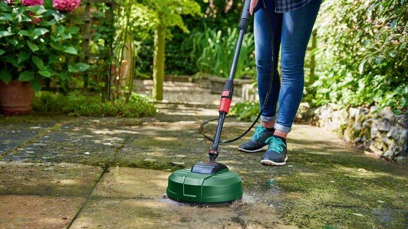 Eine Person reinigt eine Steinpflasterfläche im Garten mit einem Hochdruckreiniger. Umgeben von Pflanzen und Büschen.
