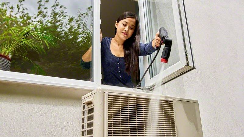 Eine Frau reinigt eine Klimaanlage, die sich außerhalb eines Fensters befindet, mit einer Bürste. Eine Pflanze steht in der Fensterbank.