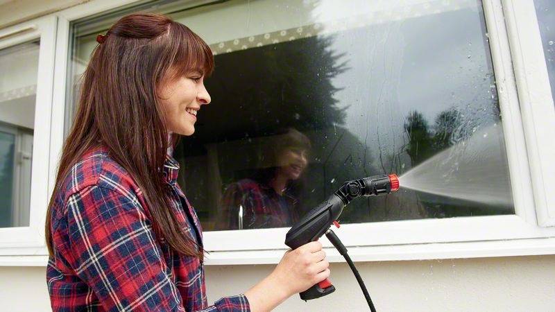 Frau reinigt ein Fenster mit einem Wasserschlauch. Sie trägt ein kariertes Hemd. Im Hintergrund ist ein Baum sichtbar.