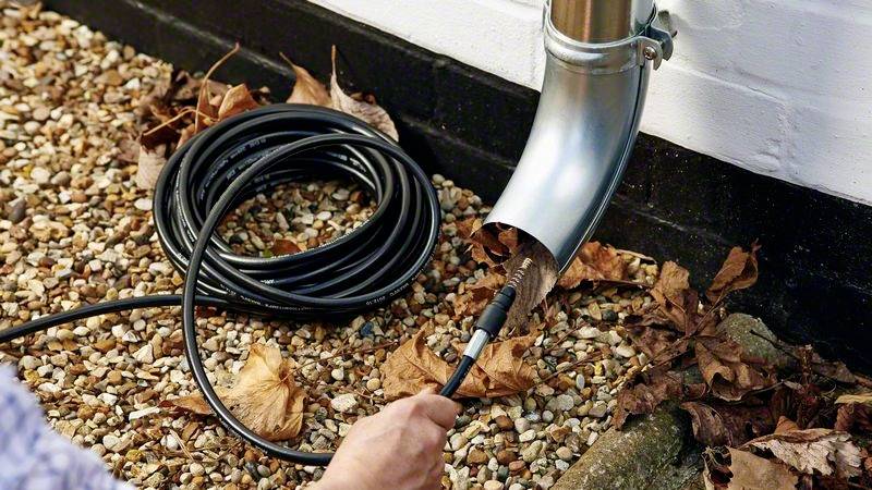 Eine Person reinigt ein Fallrohr mit einer Schlauchbürste. Herbstblätter liegen auf Kies am Boden.