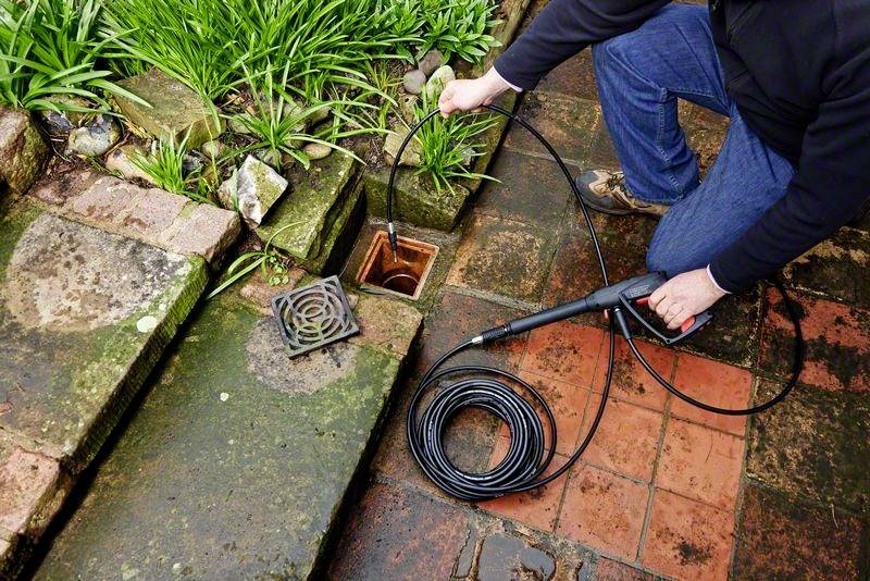 Eine Person reinigt eine verstopfte Abflussrinne im Garten mit einem Hochdruckschlauch. Pflanzen und nasses Pflaster sichtbar.