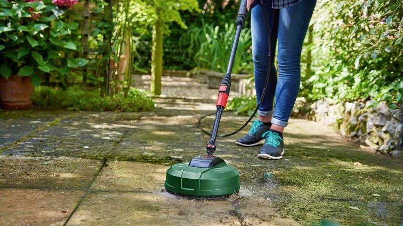 Eine Person reinigt mit einem Hochdruckreiniger eine Terrasse im Garten. Im Hintergrund sind Pflanzen und Sträucher sichtbar.