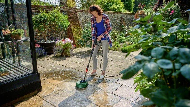 Eine Frau reinigt mit einem Hochdruckreiniger die Terrasse in einem Garten. Im Hintergrund sind Pflanzen und ein Steinzaun sichtbar.