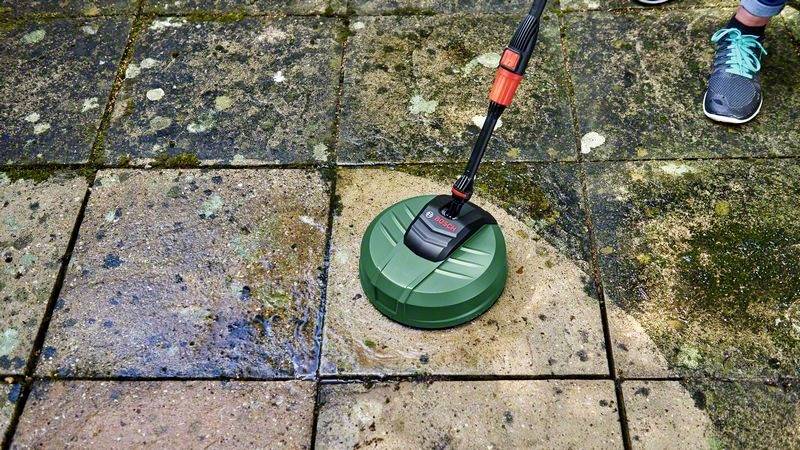 Eine Person reinigt eine stark verschmutzte Terrasse mit einem Hochdruckreiniger. Links ist der Bereich sauber, rechts noch schmutzig.