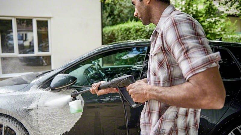 Eine Person wäscht ein schwarzes Auto mit einem Hochdruckreiniger. Das Auto ist mit weißem Schaum bedeckt. Im Hintergrund sind Bäume zu sehen.