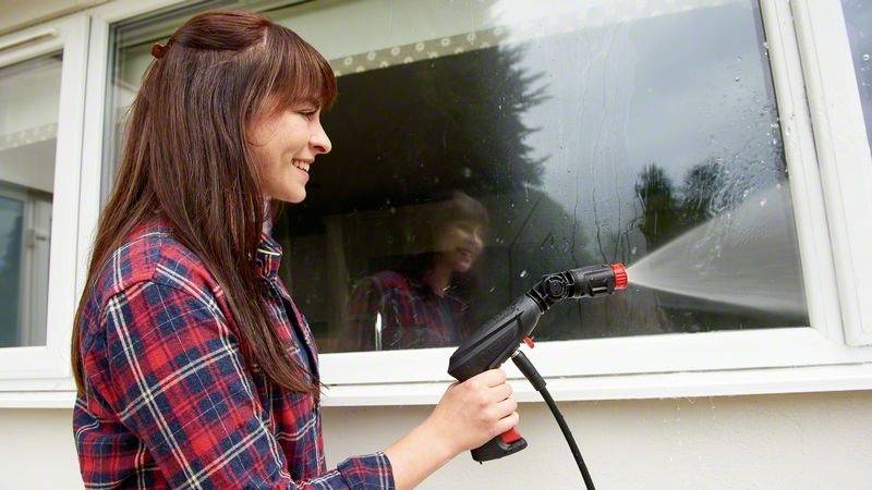 Eine Frau spritzt mit einem Gartenschlauch Wasser auf ein Fenster, das gereinigt wird. Sie trägt ein kariertes Hemd und lächelt.