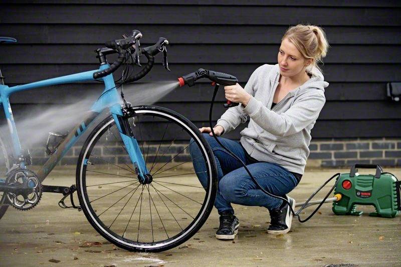 Eine Person reinigt ein blaues Fahrrad mit einem Hochdruckreiniger auf einem gepflasterten Bereich. Eine schwarze Wand ist im Hintergrund.