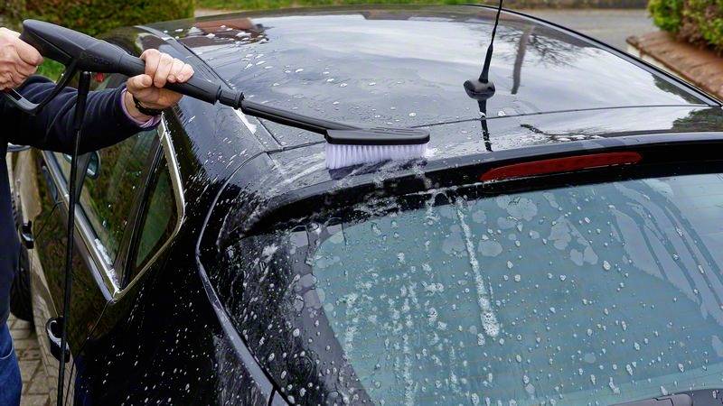 Person reinigt das Dach eines schwarzen Autos mit einer Bürste und Seifenwasser. Auto steht draußen auf einer gepflasterten Fläche.