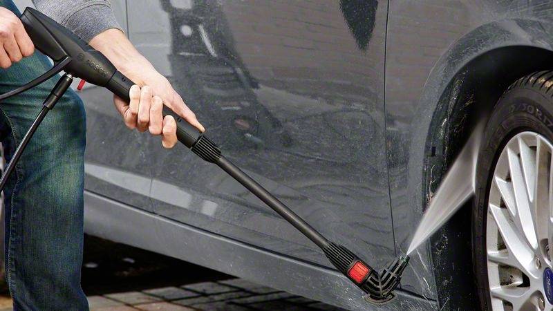 Eine Person reinigt die Seite eines Autos mit einem Hochdruckreiniger, Wasser spritzt gegen das Fahrzeug.