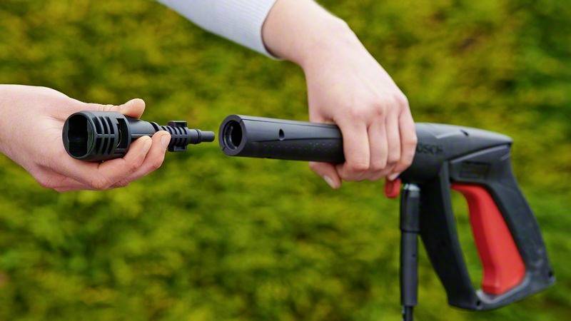 Eine Person hält einen Hochdruckreiniger und montiert eine Düse, bereit für die Reinigung. Hintergrund zeigt unscharfes Grün.