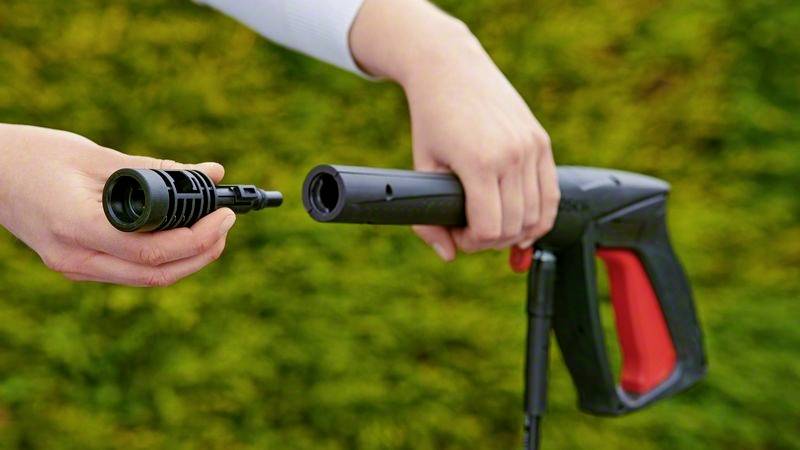 Zwei Hände montieren ein Sprühdüsenaufsatz auf einen Hochdruckreiniger vor unscharfem Hintergrund im Freien.