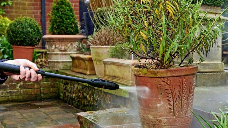 Eine Person reinigt einen Terrakotta-Pflanztopf mit einem Hochdruckreiniger in einem Garten, umgeben von mehreren Pflanzen.