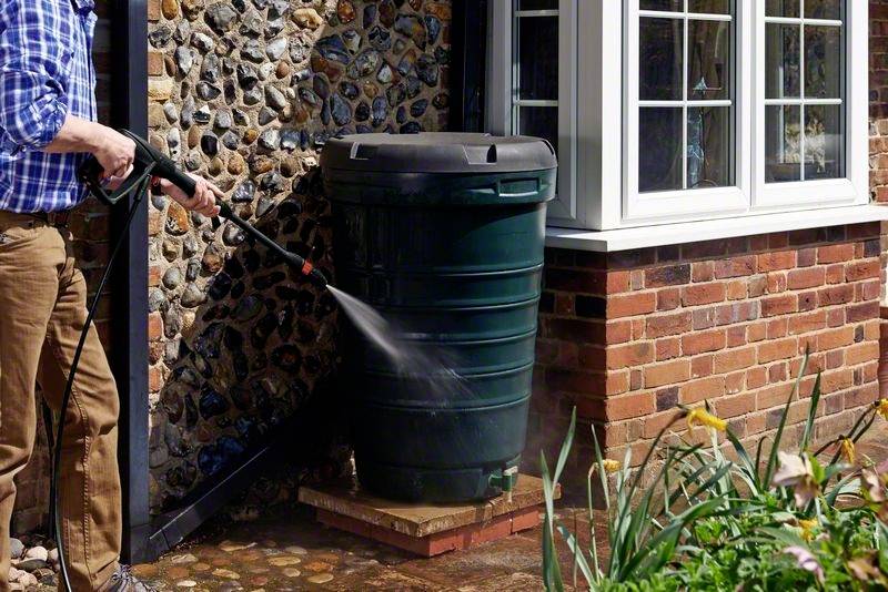 Eine Person verwendet einen Hochdruckreiniger, um ein großes grünes Regentonnenfass neben einem Ziegelhaus zu reinigen.