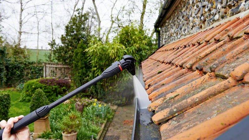 Person reinigt Dachrinne eines Hauses mit Hochdruckreiniger; Wasser entfernt Moos von Ziegeln. Garten im Hintergrund sichtbar.